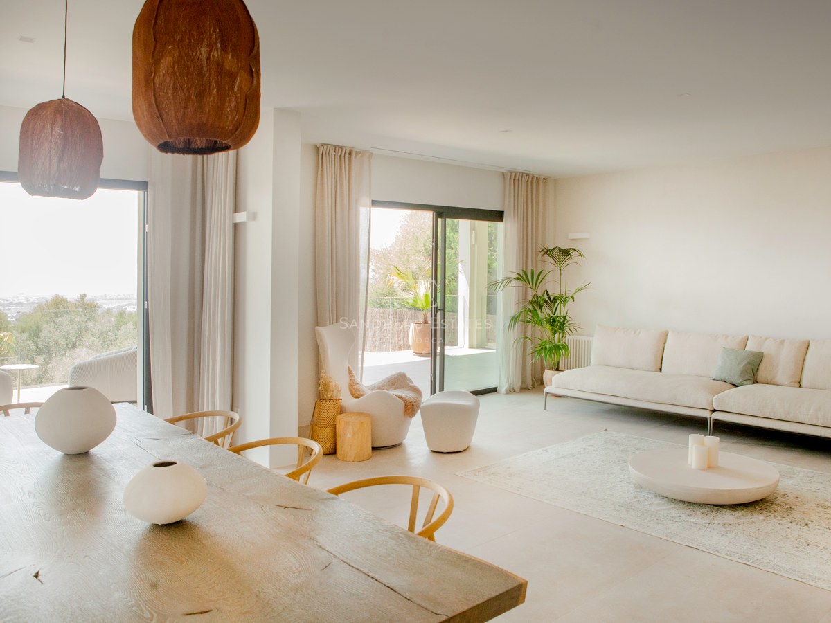 Helles modernes Wohnzimmer mit großem Holzesstisch, weißem Sofa und bodentiefen Fenstern zur Terrasse.