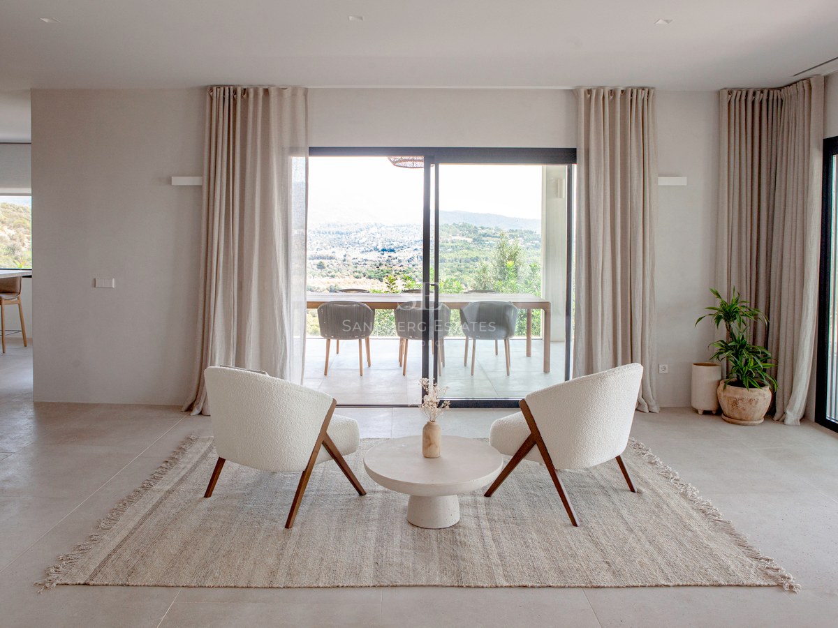 Modernes Wohnzimmer mit zwei cremefarbenen Sesseln, Juteteppich und bodentiefen Fenstern mit Blick auf die Berge.