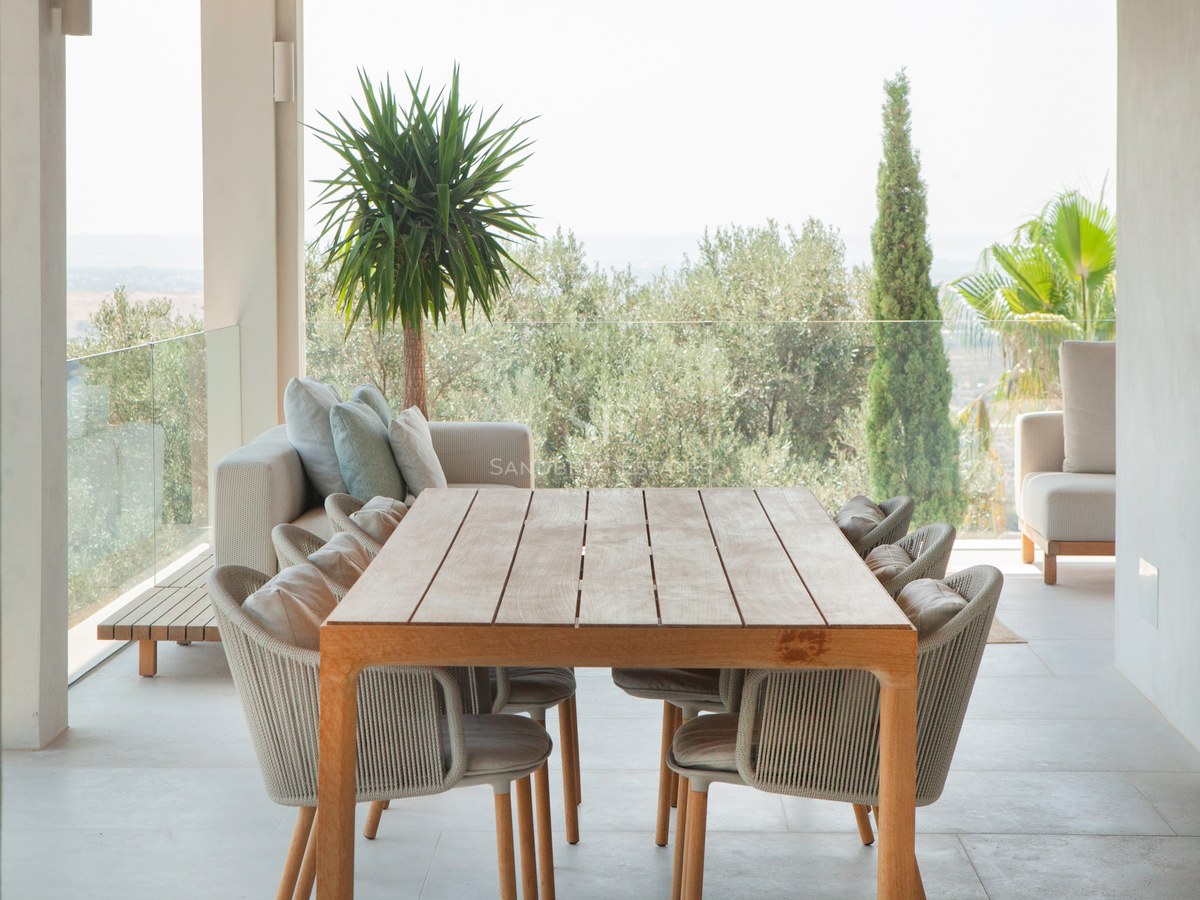 Moderne überdachte Terrasse mit großem Holztisch, Korbstühlen und Blick auf mediterrane Vegetation.