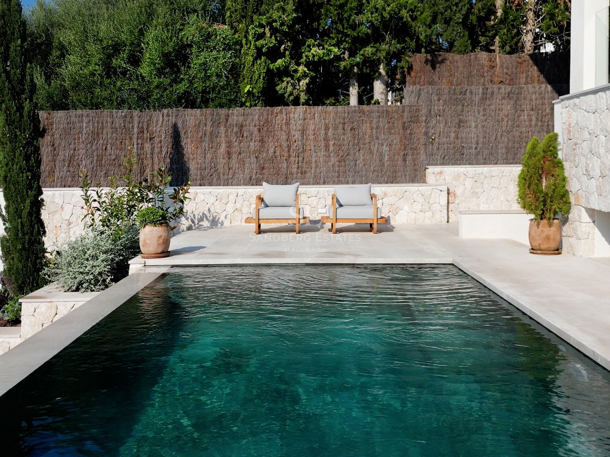 Türkisfarbener Pool neben weißen Natursteinmauern mit Holzliegen und mediterranen Pflanzen im Hintergrund.