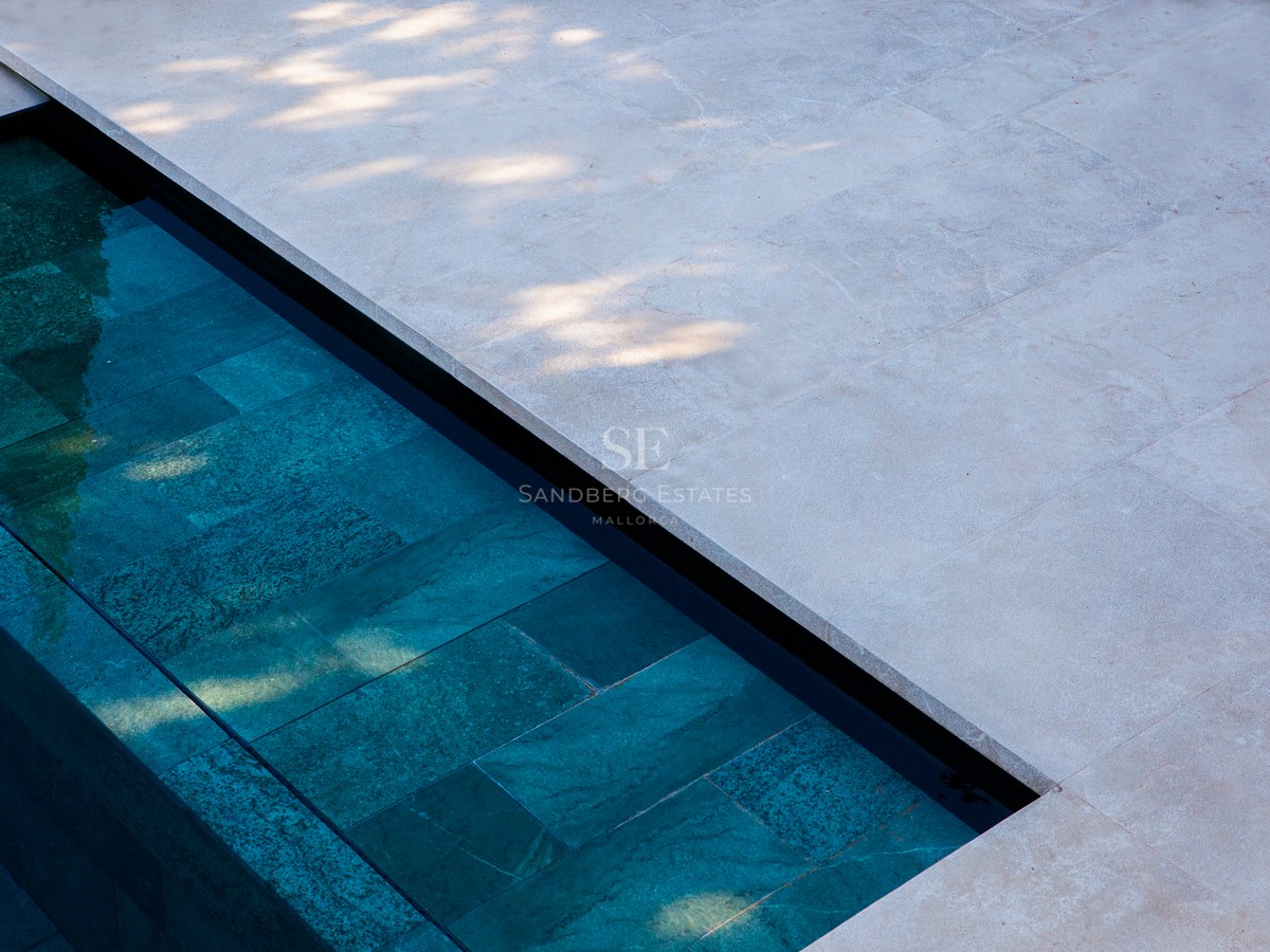 Nahaufnahme eines modernen Pools mit türkisblauen Steinfliesen und hellem, sandfarbenem Naturstein-Terrassenbelag.