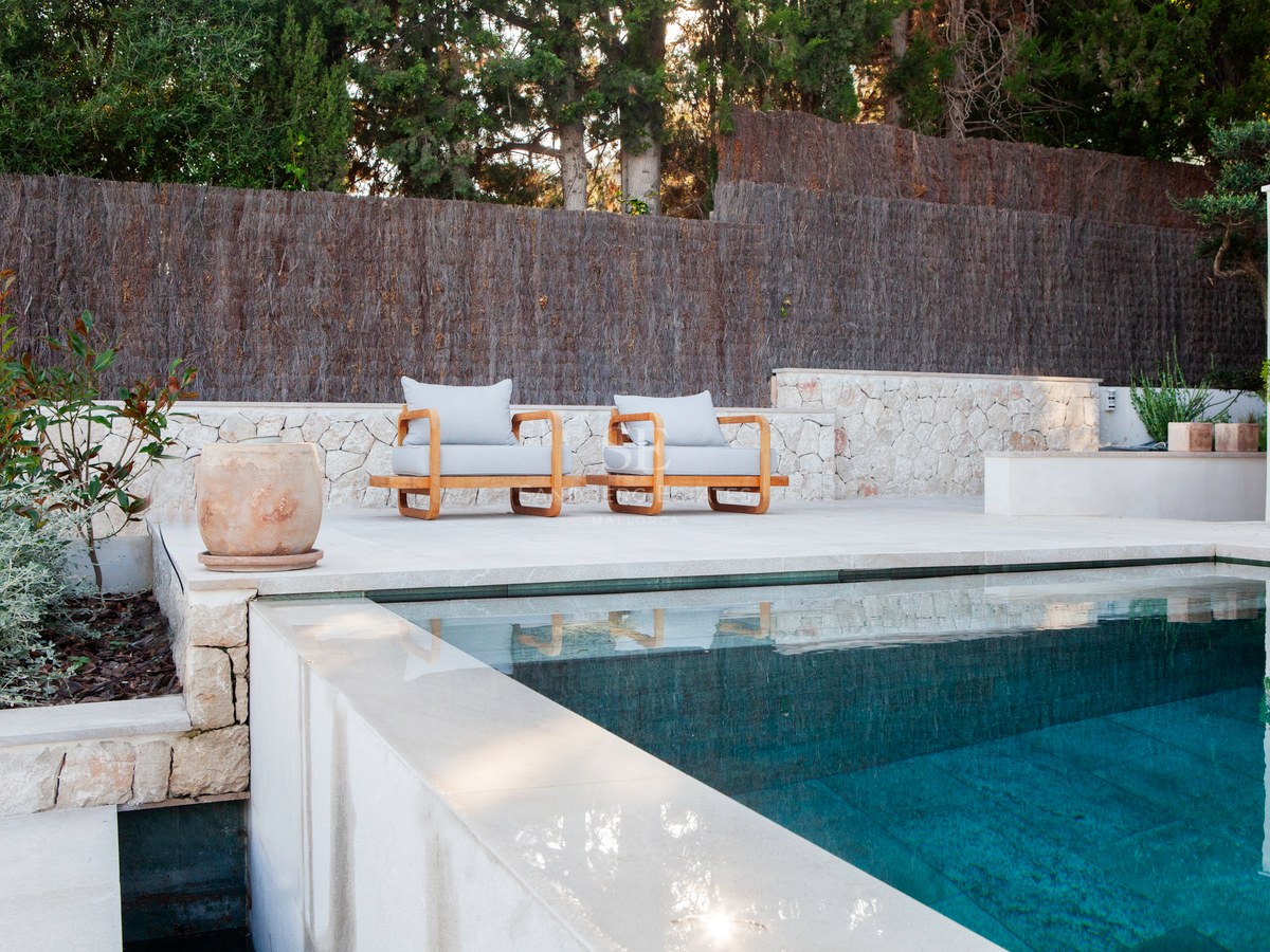 Infinity-Pool mit türkisblauem Wasser neben einer Kalksteinterrasse mit zwei Designer-Holzsesseln.