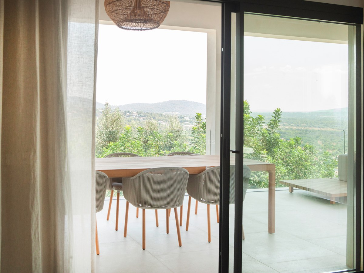 Blick durch Glastüren auf eine überdachte Terrasse mit Holztisch, Korbstühlen und Berglandschaft.