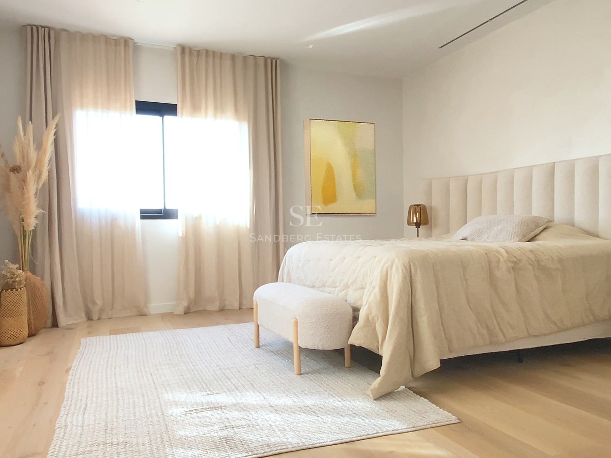 Helles Hauptschlafzimmer mit hellem Holzboden, raumhohen Vorhängen und gepolstertem Kopfteil in neutralen Tönen.