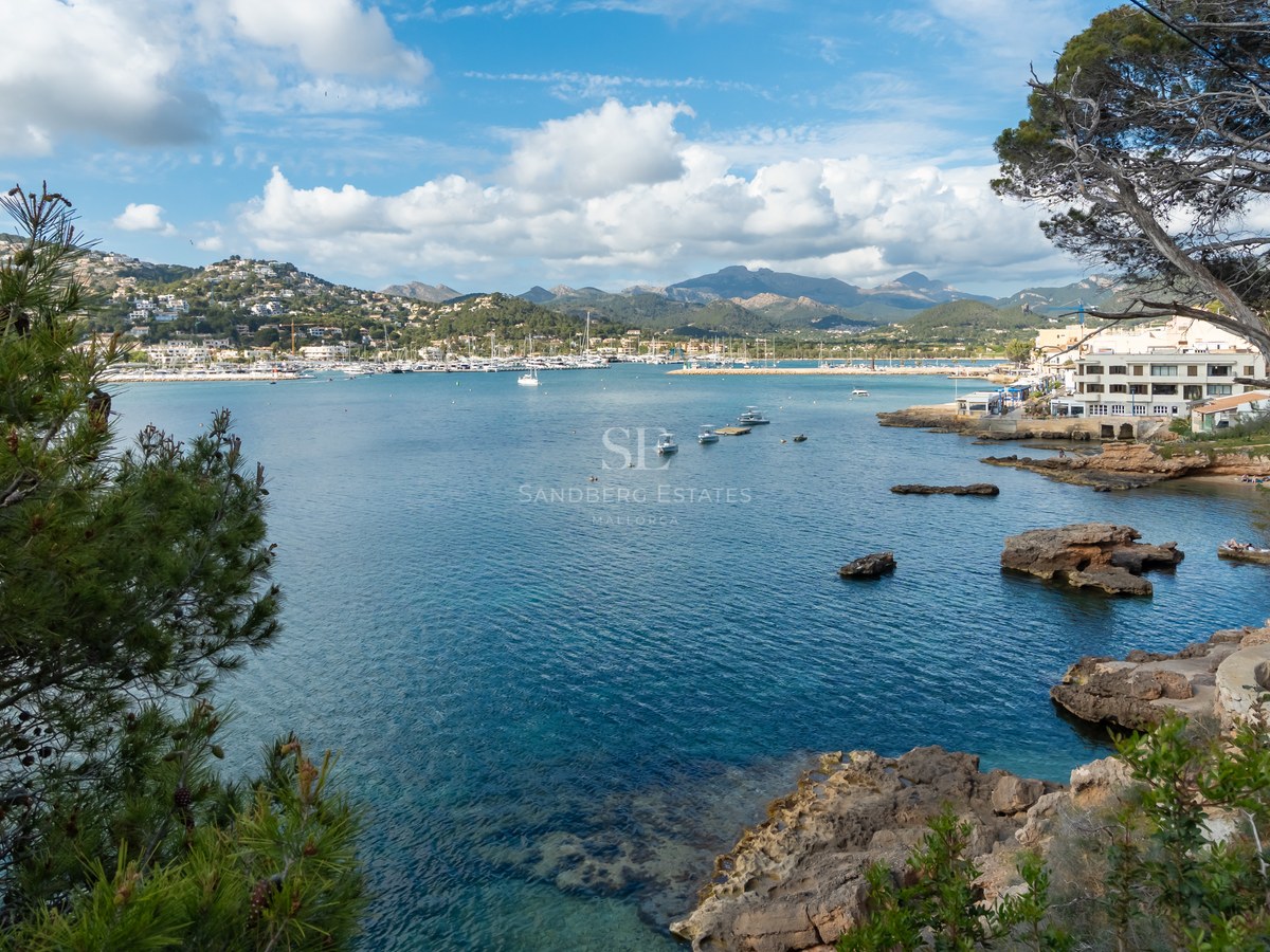 Vista elevada de una bahía mediterránea con yates, costa rocosa y montañas bajo un cielo azul con nubes.