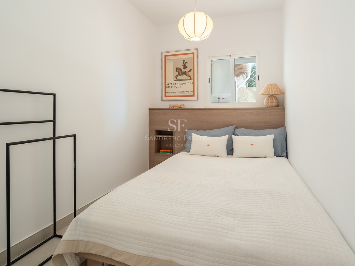 Dormitorio minimalista con cama blanca, cabecero de madera clara, ventana y lámpara de papel colgante.