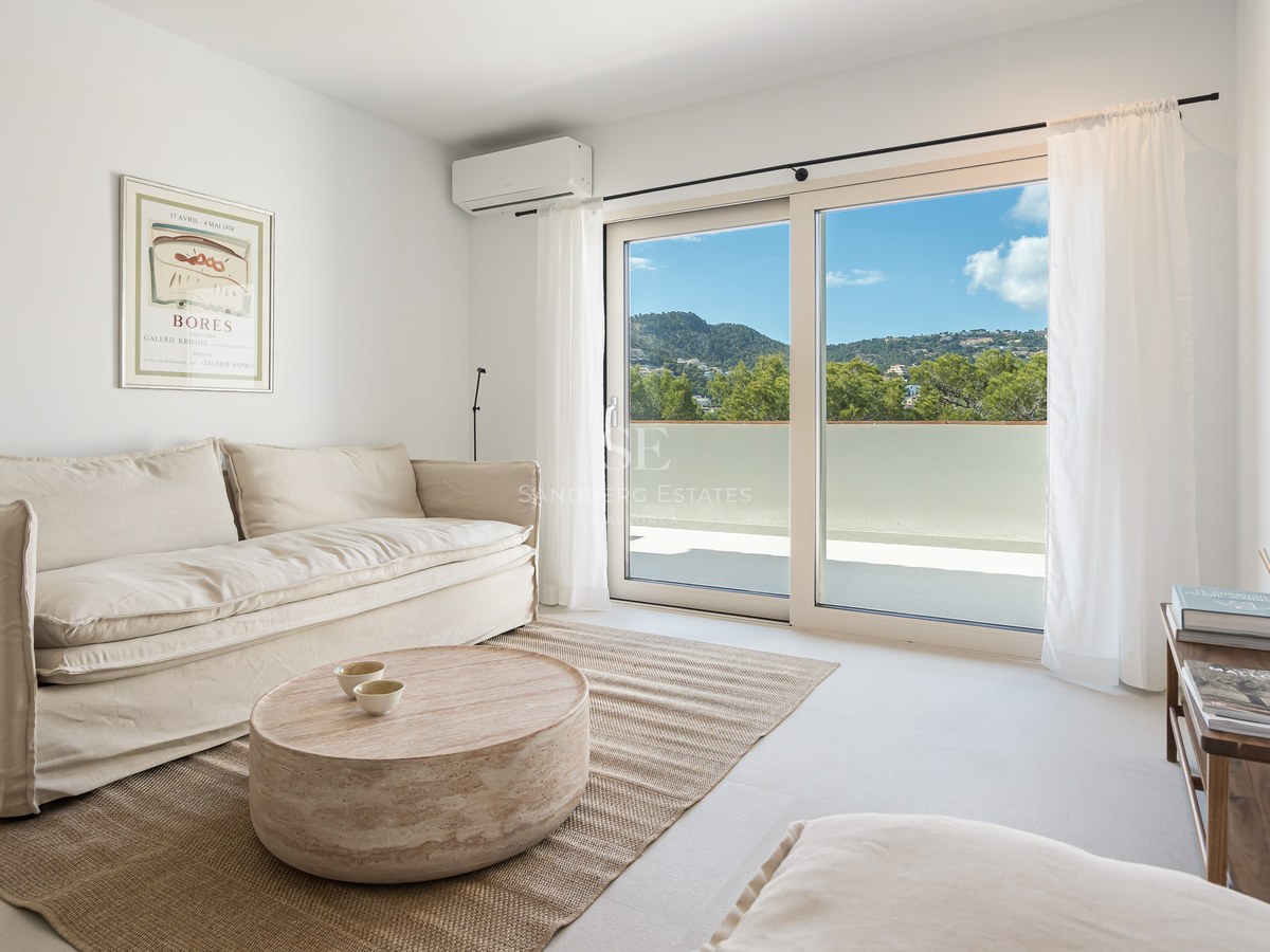 Salón moderno con sofá de lino blanco y grandes ventanales con vistas a la vegetación de montaña.