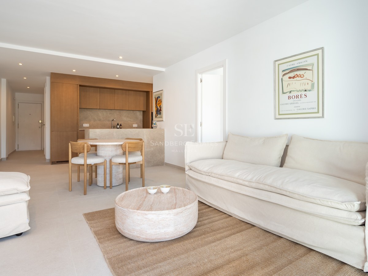 Salón luminoso con sofá de lino blanco, mesa de centro de madera redonda y cocina integrada al fondo.