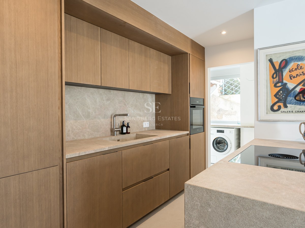 Cocina moderna con armarios de roble sin tiradores, encimera de piedra, horno integrado y zona de lavandería al fondo.