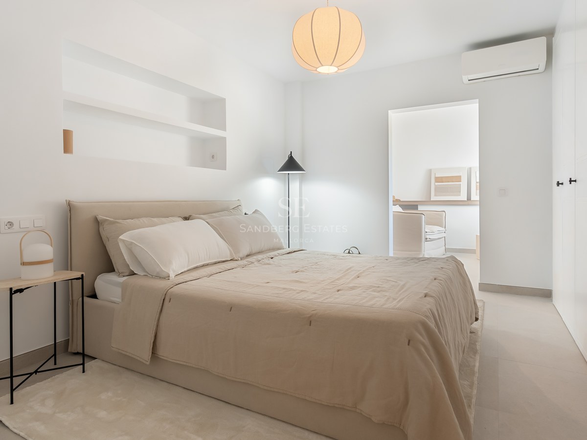Dormitorio moderno con cama beige, estanterías integradas en la pared, lámpara de papel redonda y suelos claros.