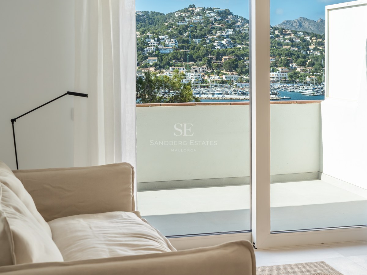 Interior con sofá beige junto a ventanales de suelo a techo con vistas a un pueblo costero y puerto deportivo.