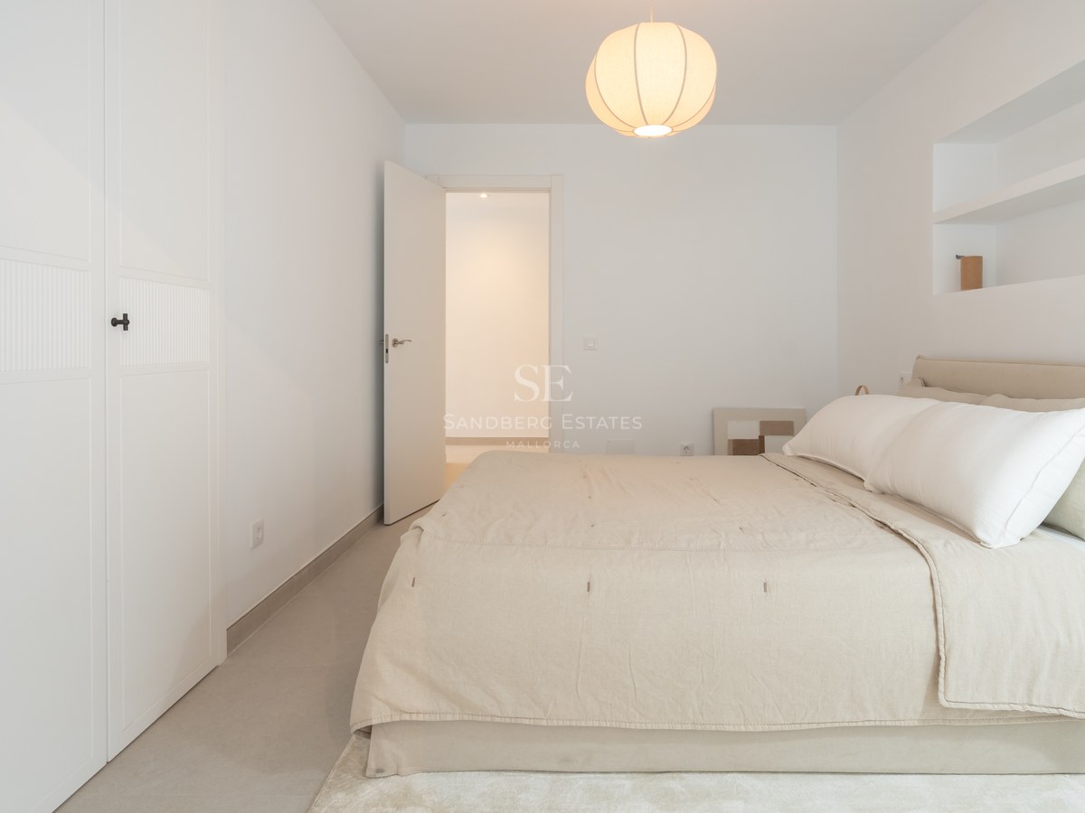 Un dormitorio luminoso y minimalista con una cama grande, sábanas blancas, una lámpara colgante redonda y una puerta abierta.
