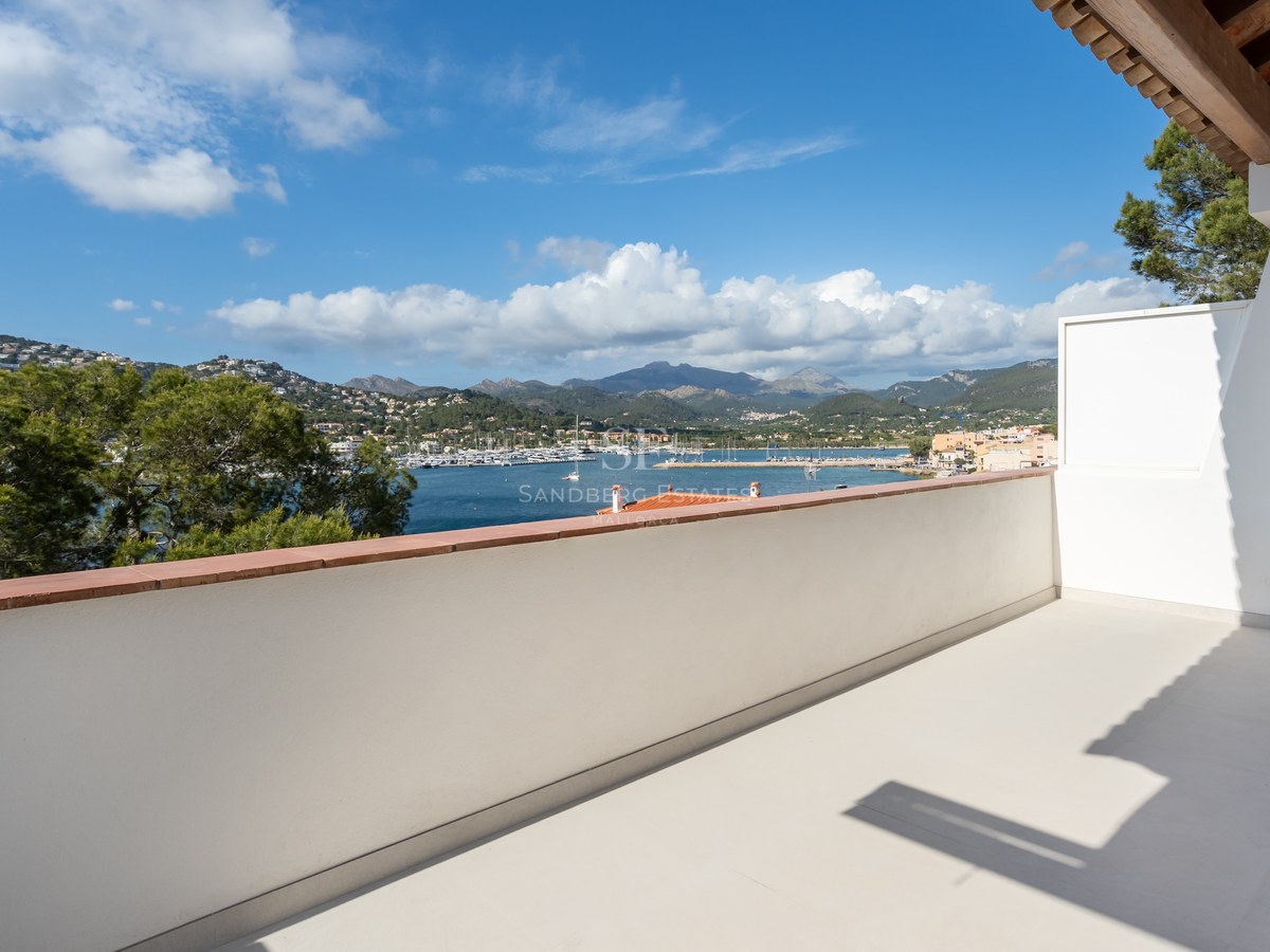 Amplia terraza blanca con vistas a un puerto deportivo con veleros, rodeada de colinas verdes y montañas.