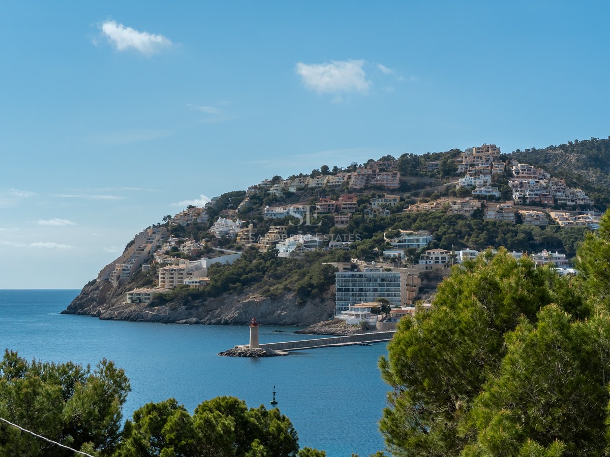 Paisaje mediterráneo con villas en la colina frente a una bahía azul con faro y pinos en primer plano.