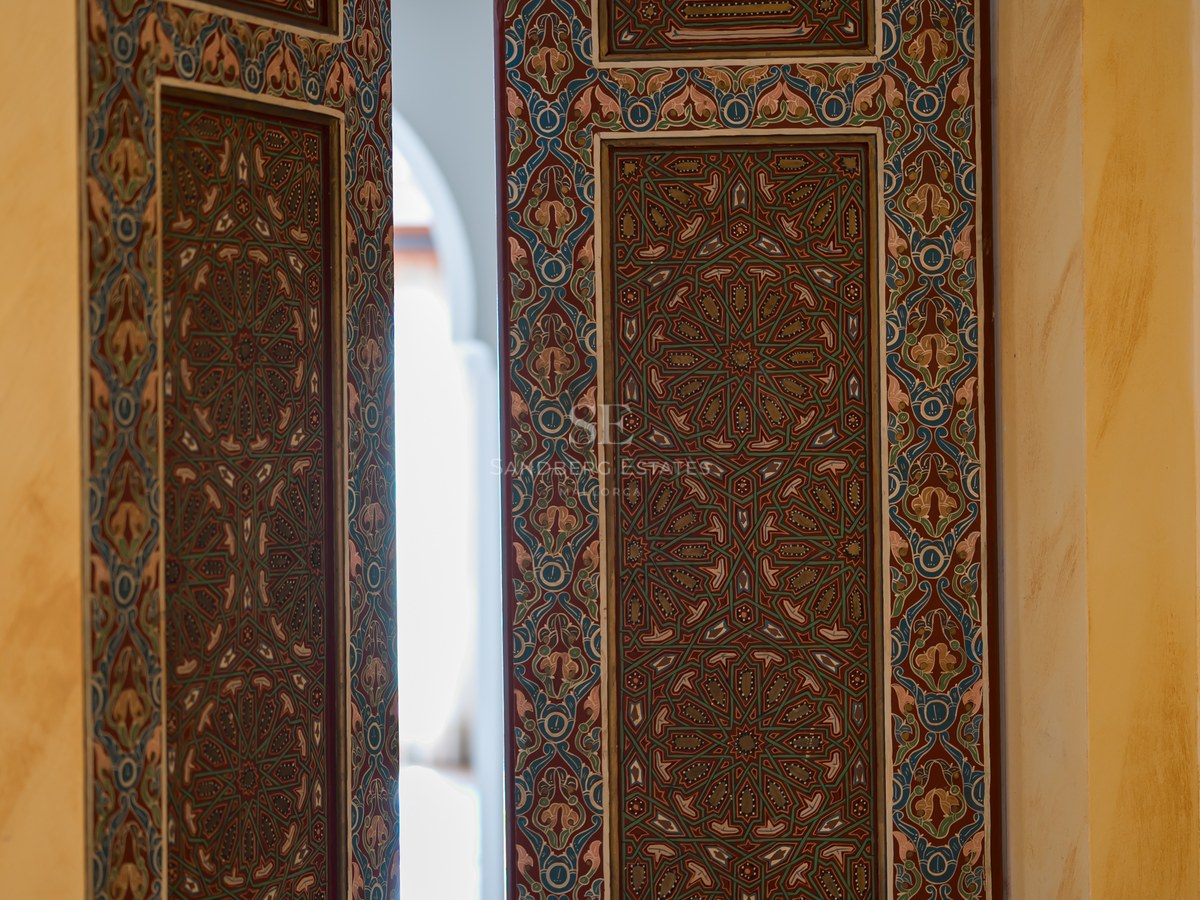 Motifs mauresques complexes sur une porte en bois sombre s'ouvrant sur un patio lumineux.