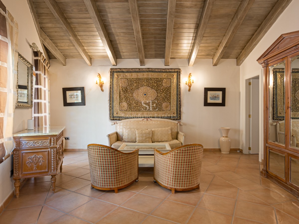 Espace de vie avec sol en terre cuite, plafond en poutres de bois, fauteuils en osier et tapisserie murale.