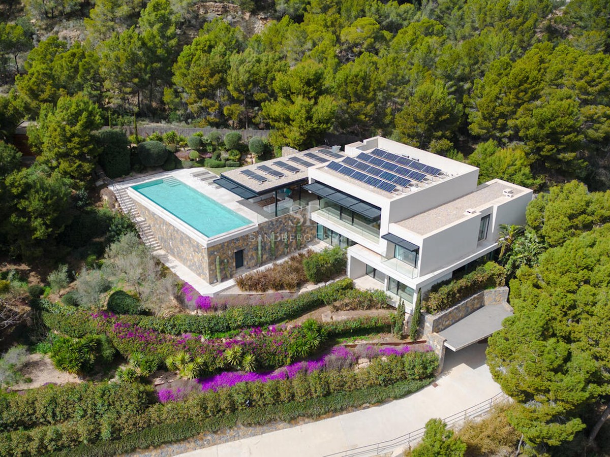 Luftaufnahme einer modernen weißen Villa mit Infinity-Pool, Sonnenkollektoren und umgebendem Pinienwald.