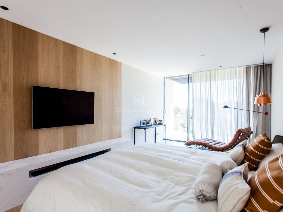 Modernes Schlafzimmer mit Eichenholz-TV-Wand, weißem Marmorsockel und Leder-Loungesessel vor Terrassentür.