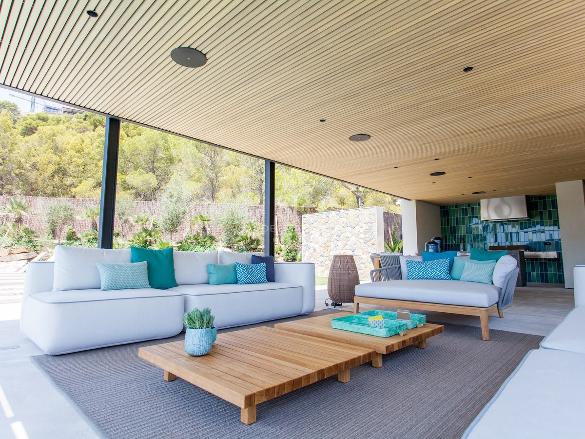 Moderne überdachte Terrasse mit weißen Sofas, türkisfarbenen Kissen, Holztischen und einer Sommerküche.