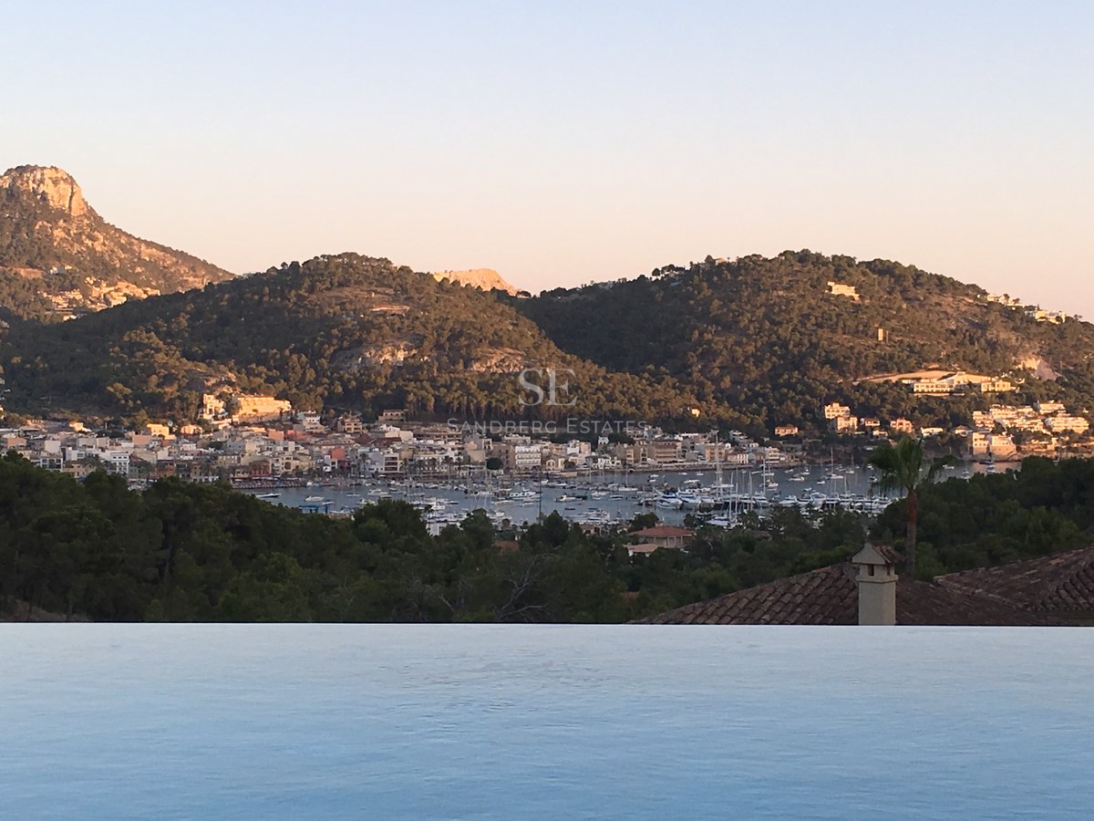 Rand eines Infinity-Pools mit Blick auf einen mediterranen Hafen mit Booten und grünen Bergen bei Sonnenuntergang.