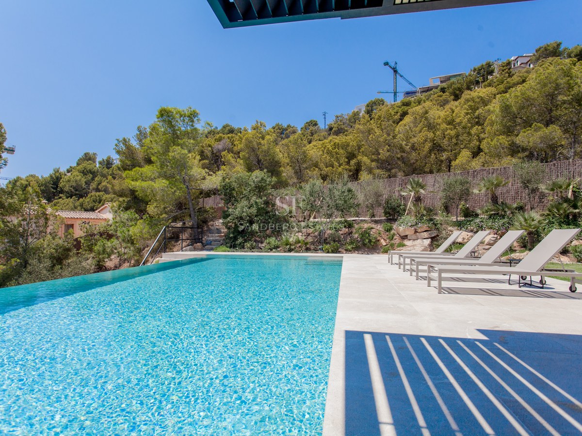 Großer Infinity-Pool mit türkisblauem Wasser und Steinterrasse mit grauen Liegen vor einem bewaldeten Hügel.