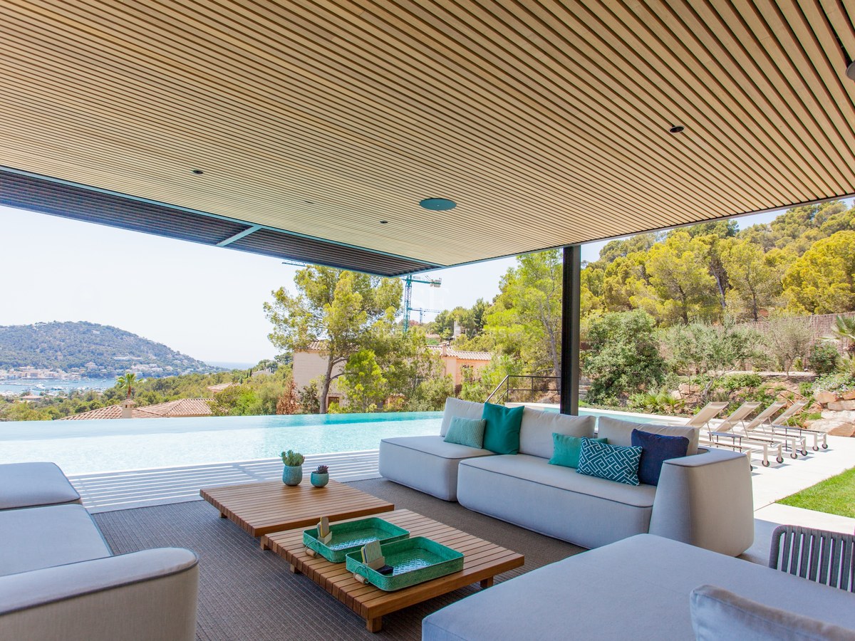 Moderne Terrasse mit grauen Sofas, Holzlattenendecke und Infinity-Pool mit Blick auf Küste und Berge.
