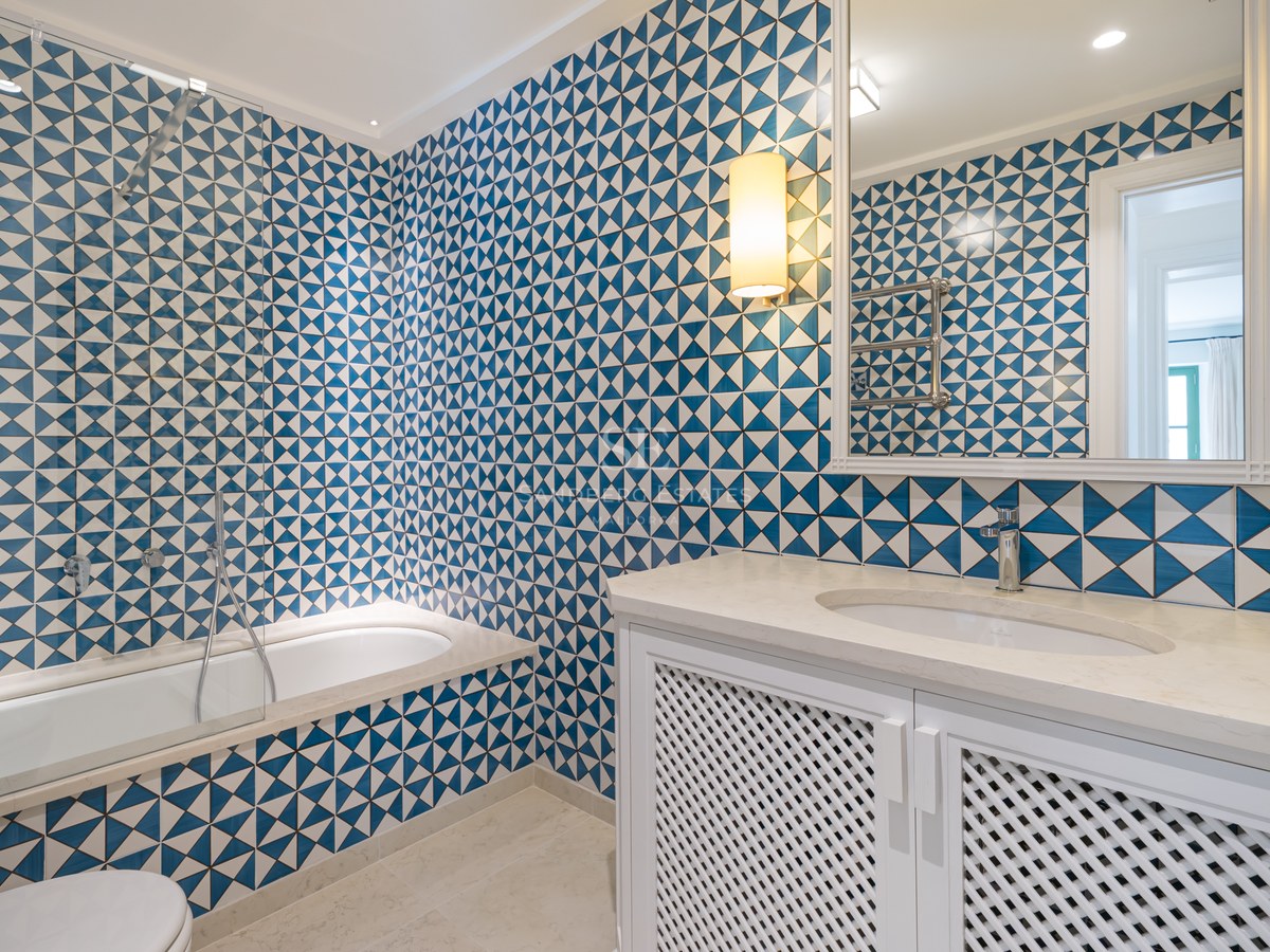 Baño con azulejos geométricos en azul y blanco, lavabo blanco con puertas de celosía y bañera integrada.