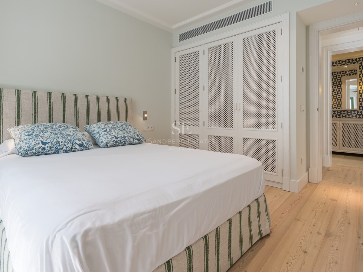 Dormitorio luminoso con cama blanca, cabecero de rayas verdes, suelos de madera clara y armarios empotrados blancos.