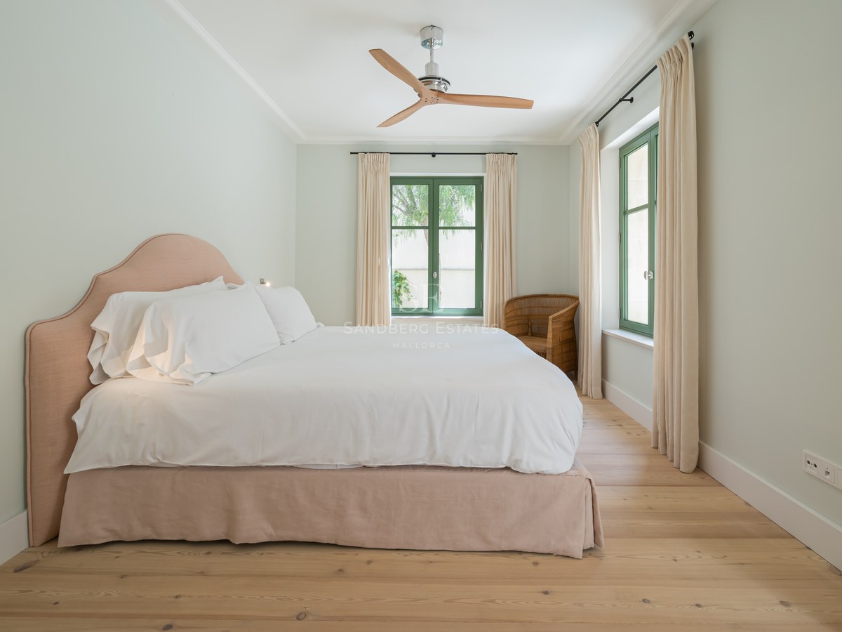 Dormitorio con cama blanca, cabecero rosa suave, ventilador de madera y ventanas de marco verde sobre suelo de roble.