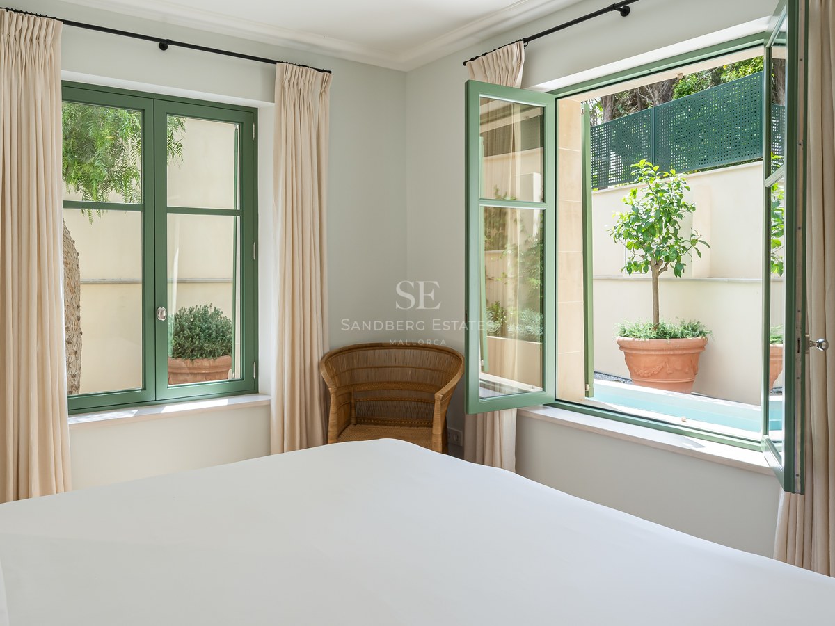 Dormitorio con cama blanca, ventanas de marco verde, sillón de mimbre y vistas a un patio con plantas.