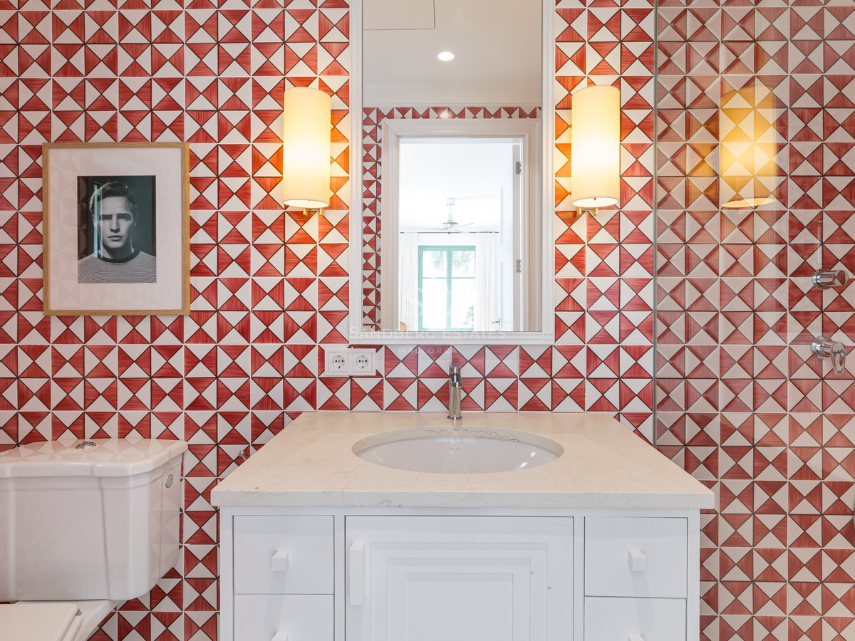 Lavabo blanco con pared de azulejos geométricos rojos y blancos, espejo y apliques de pared.