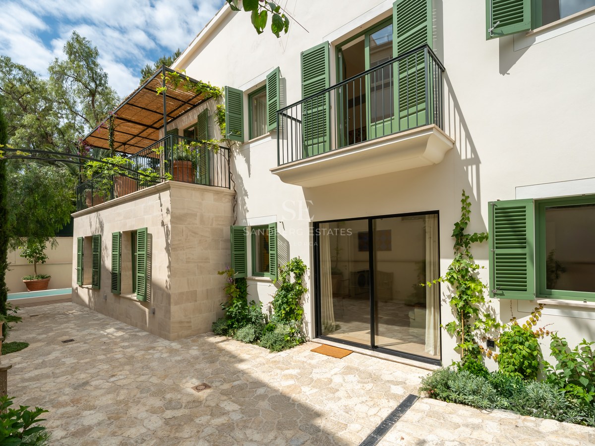 Fachada de villa blanca con persianas verdes, balcones de forja y patio de piedra natural bajo el sol.