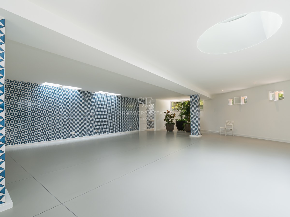 Amplia sala vacía con suelos de gres gris, pared de azulejos con motivos azules y plantas ornamentales.