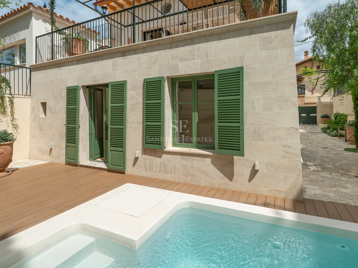 Piscina de inmersión moderna con tarima de madera junto a una fachada de piedra con contraventanas verdes.