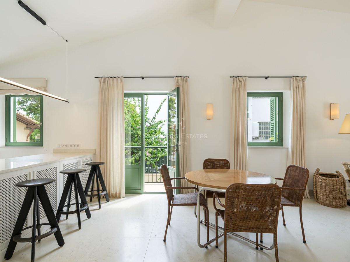 Comedor moderno con mesa redonda de madera, sillas de mimbre y puertas francesas verdes hacia un jardín frondoso.