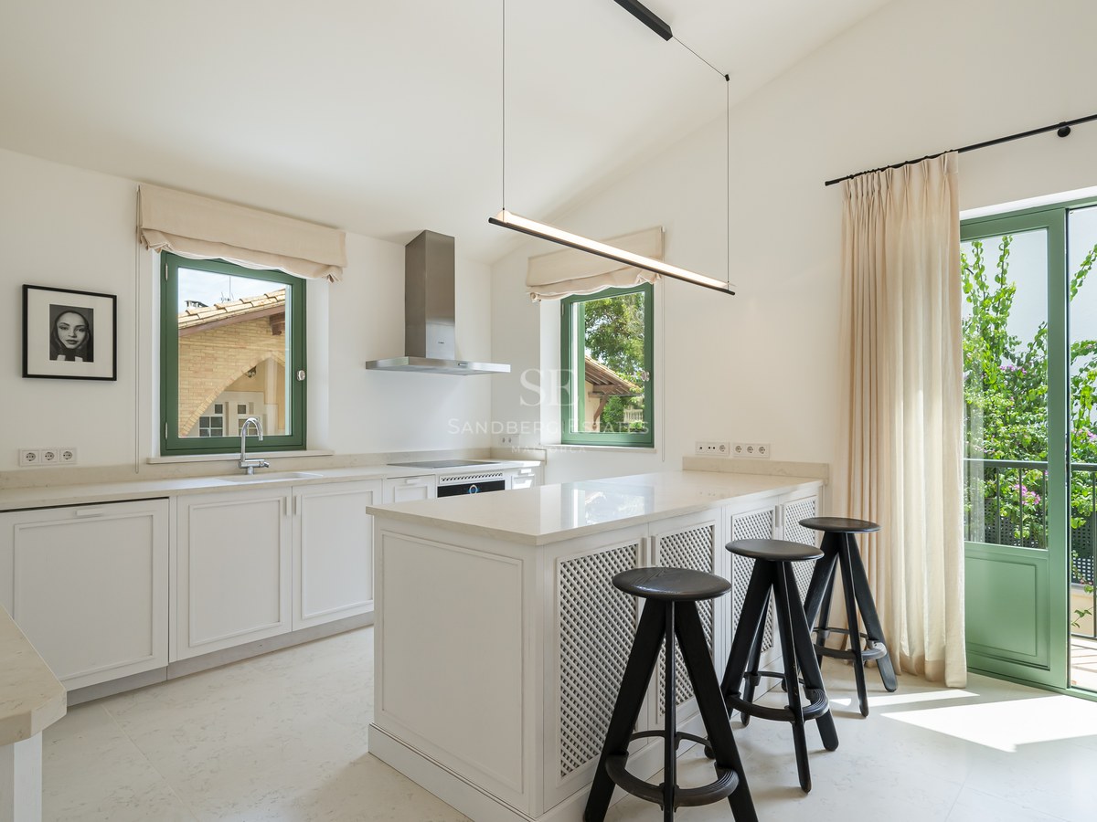 Cocina moderna blanca con isla central, taburetes negros y ventanas de marco verde inundadas de luz natural.