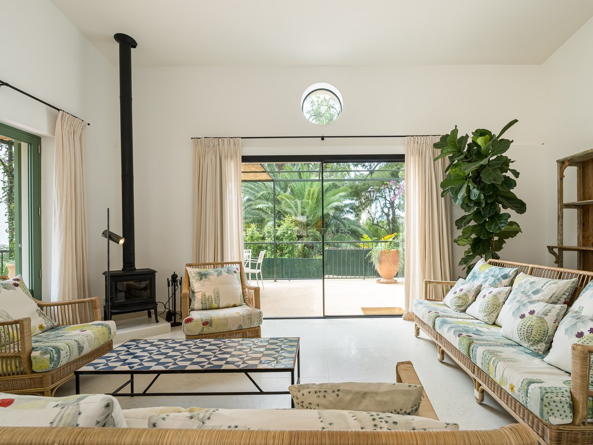 Salón soleado con muebles de bambú, estufa de leña negra y grandes puertas de cristal hacia un jardín verde.