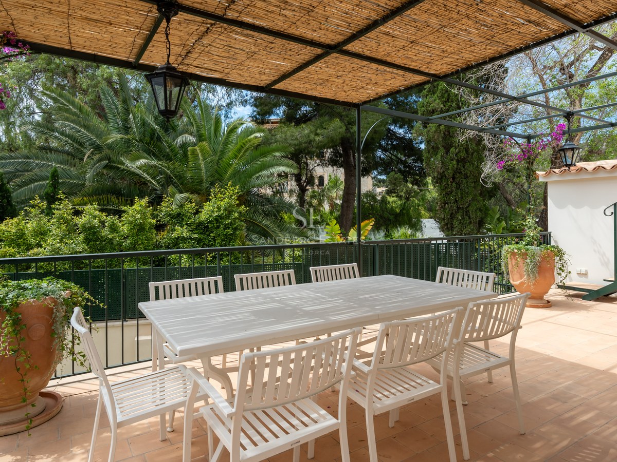 Mesa de comedor exterior blanca y sillas bajo una pérgola de cañizo con vistas a un jardín de palmeras.