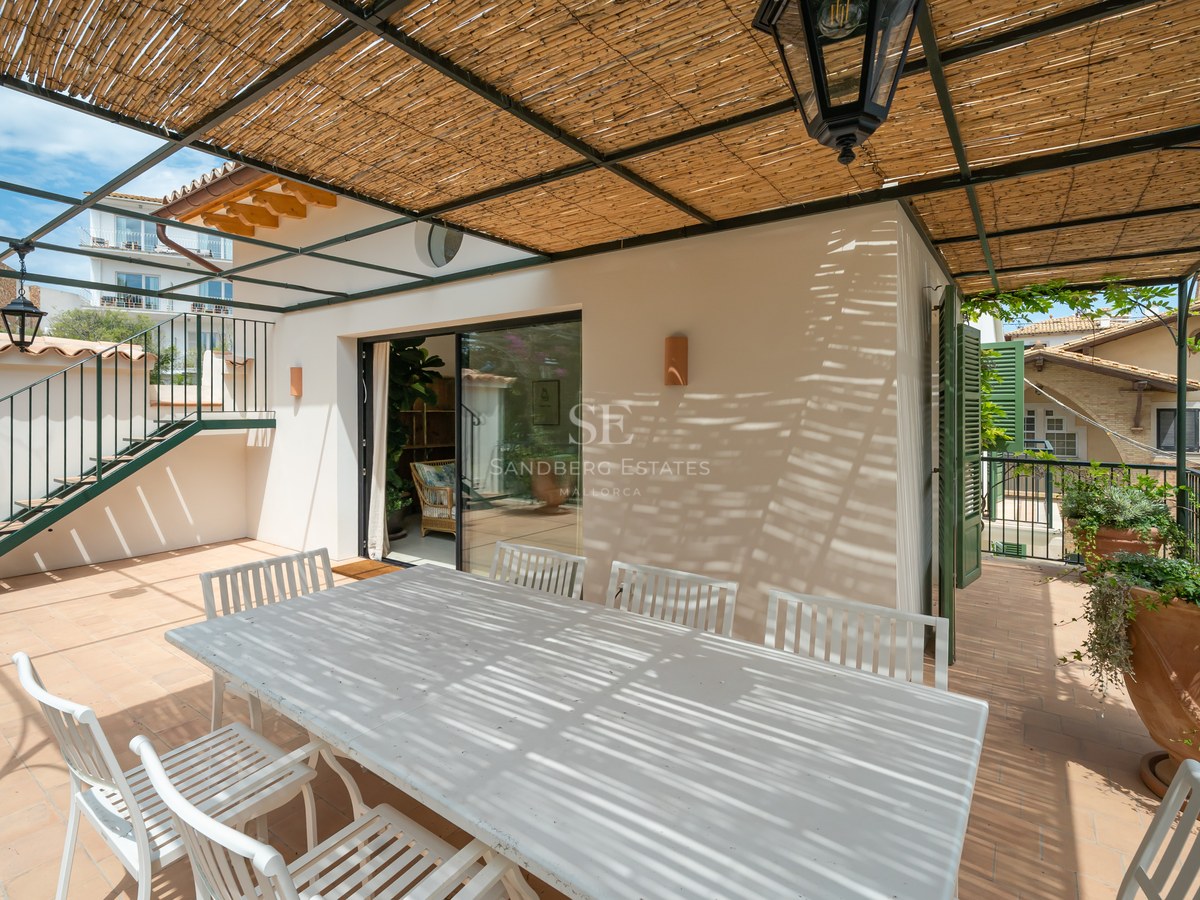 Mesa y sillas de madera blanca bajo una pérgola de cañizo con un farol tradicional en una terraza soleada.