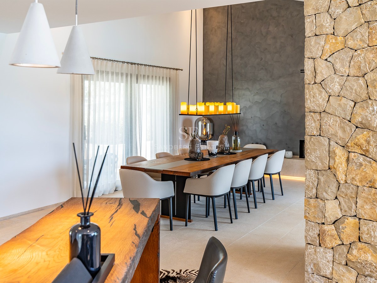 Modern matplats med långt massivt träbord, grå stolar, naturstensvägg och designad taklampa.