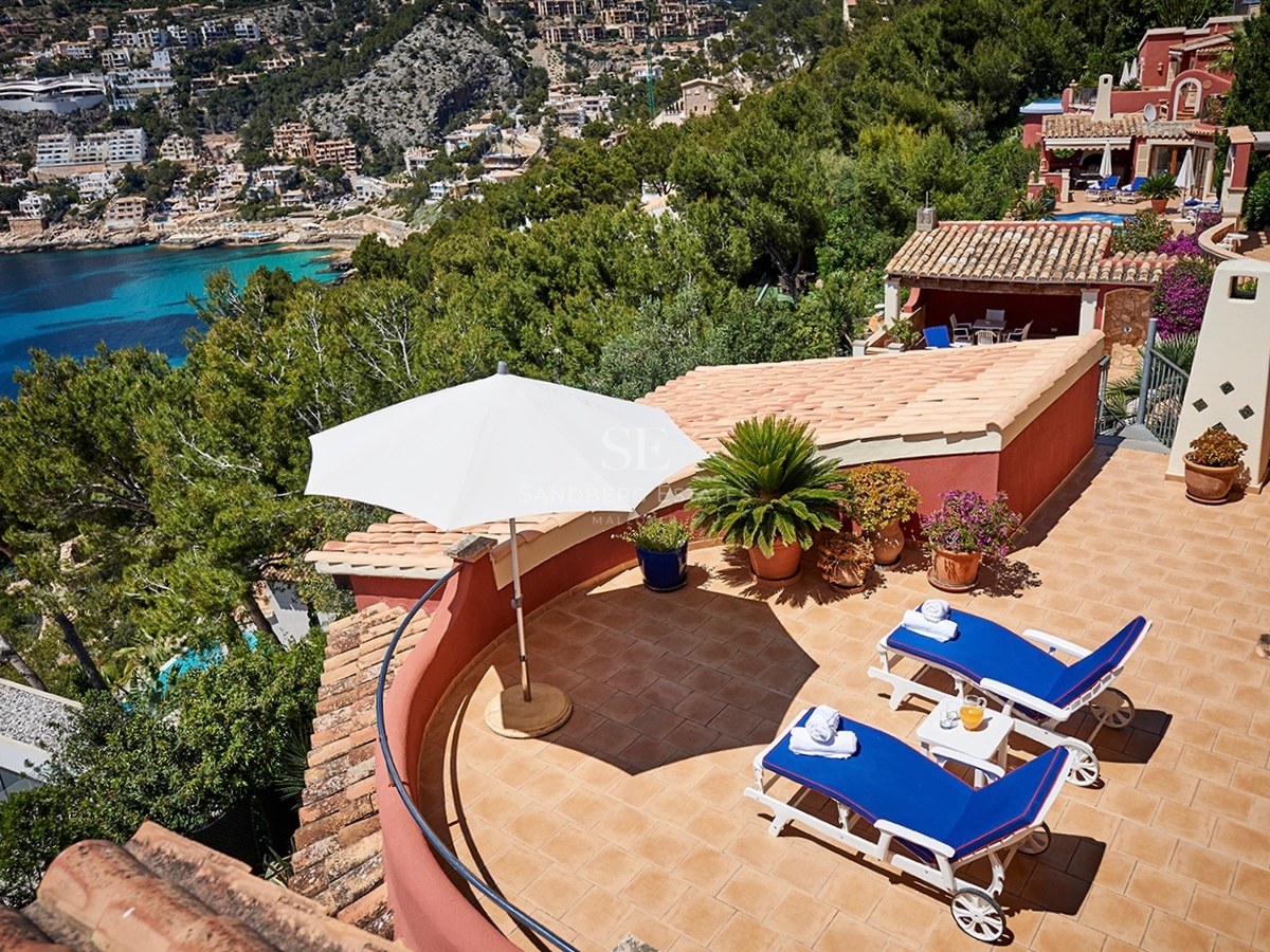 Sonnige Terrasse mit blauen Sonnenliegen, weißem Sonnenschirm und Panoramablick auf eine mediterrane Bucht.