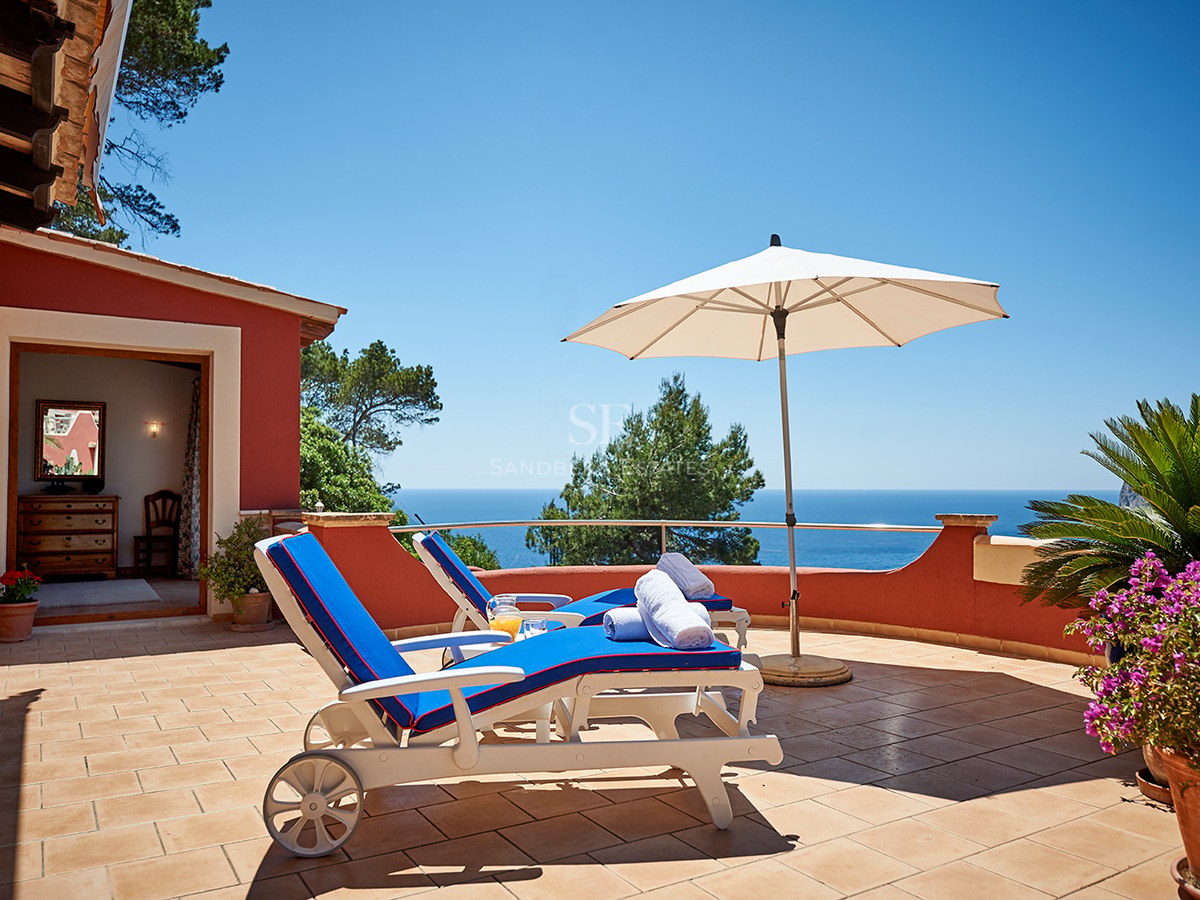 Zwei blaue Sonnenliegen auf einer Terrakotta-Terrasse mit Blick auf das strahlend blaue Meer unter klarem Himmel.