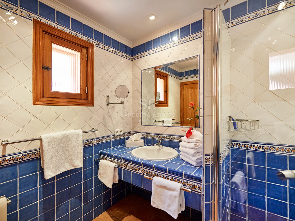 Badezimmer mit blau-weißen Fliesen, Waschbecken, Spiegel, Glasdusche und Holzfenster.