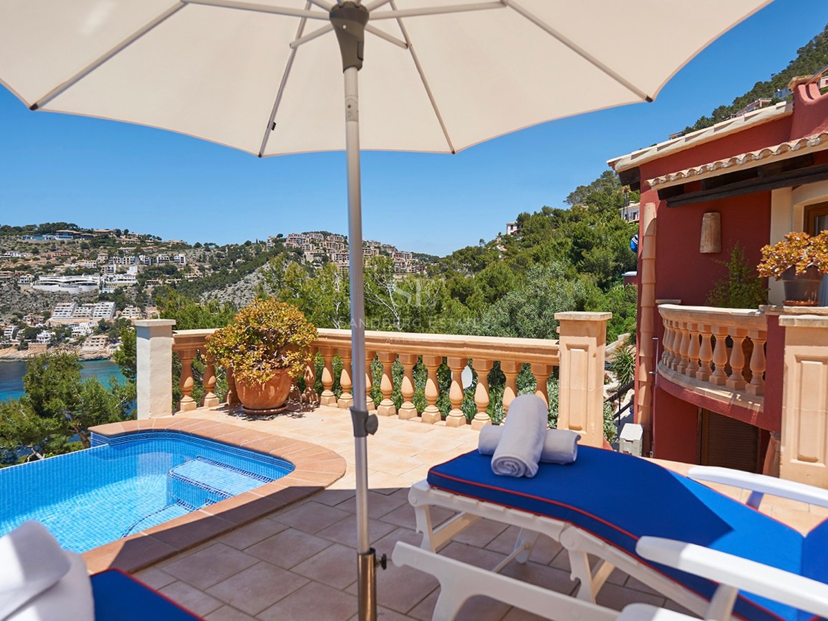 Sonnige Terrasse mit kleinem Pool, blauen Liegen und Sonnenschirm mit Blick auf eine Bucht und Hügel.