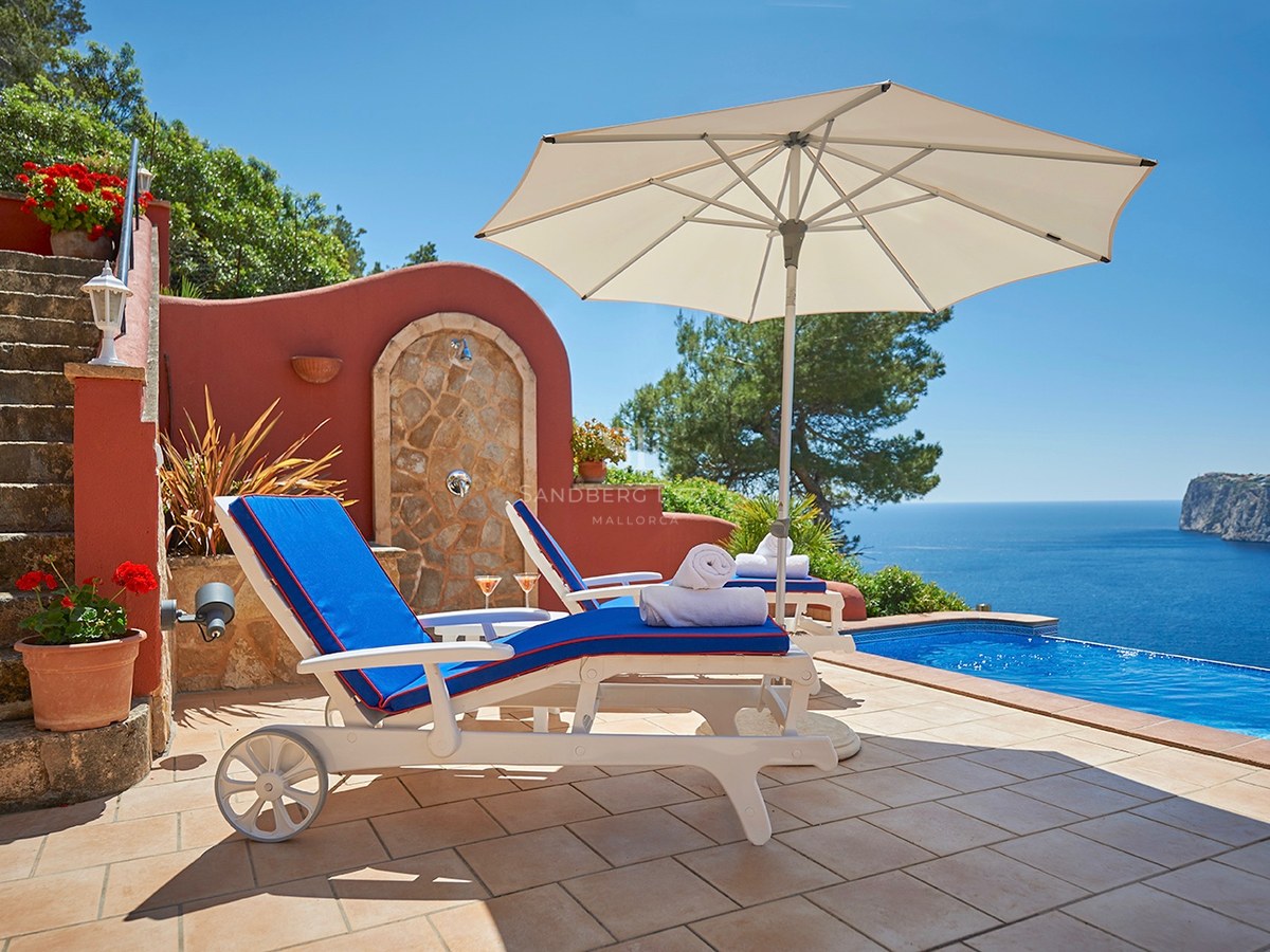 Weiße Sonnenliegen mit blauen Kissen an einem Infinity-Pool mit Blick auf das Mittelmeer und steile Klippen.