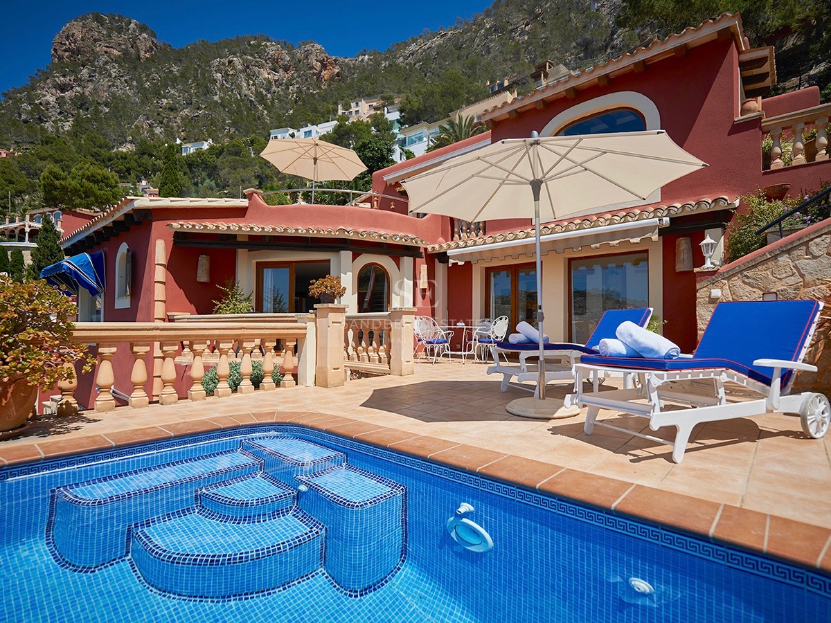 Blauer Pool und Sonnendeck mit Liegen vor einer terrakottafarbenen Villa mit Bergen im Hintergrund.
