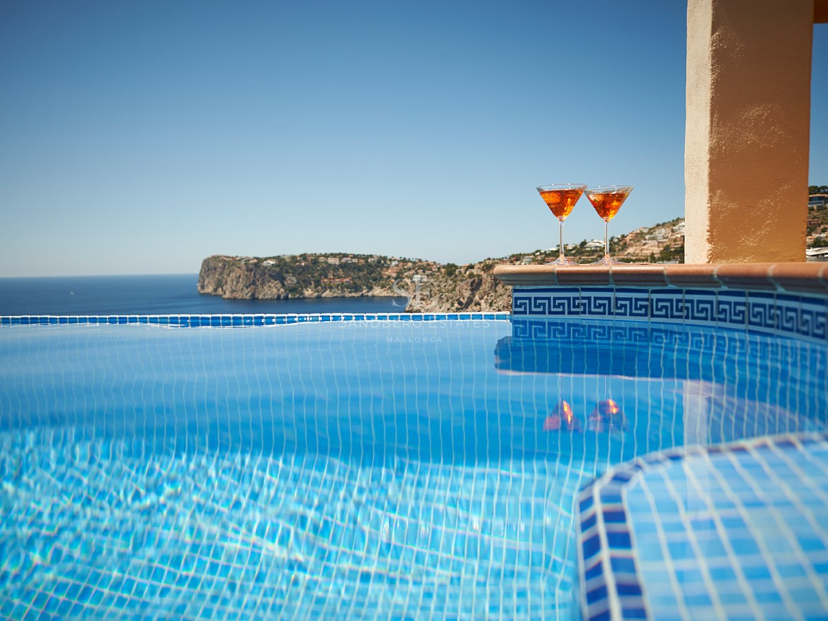 Nahaufnahme eines blau gefliesten Infinity-Pools mit zwei Cocktails am Rand und Blick auf das Meer.