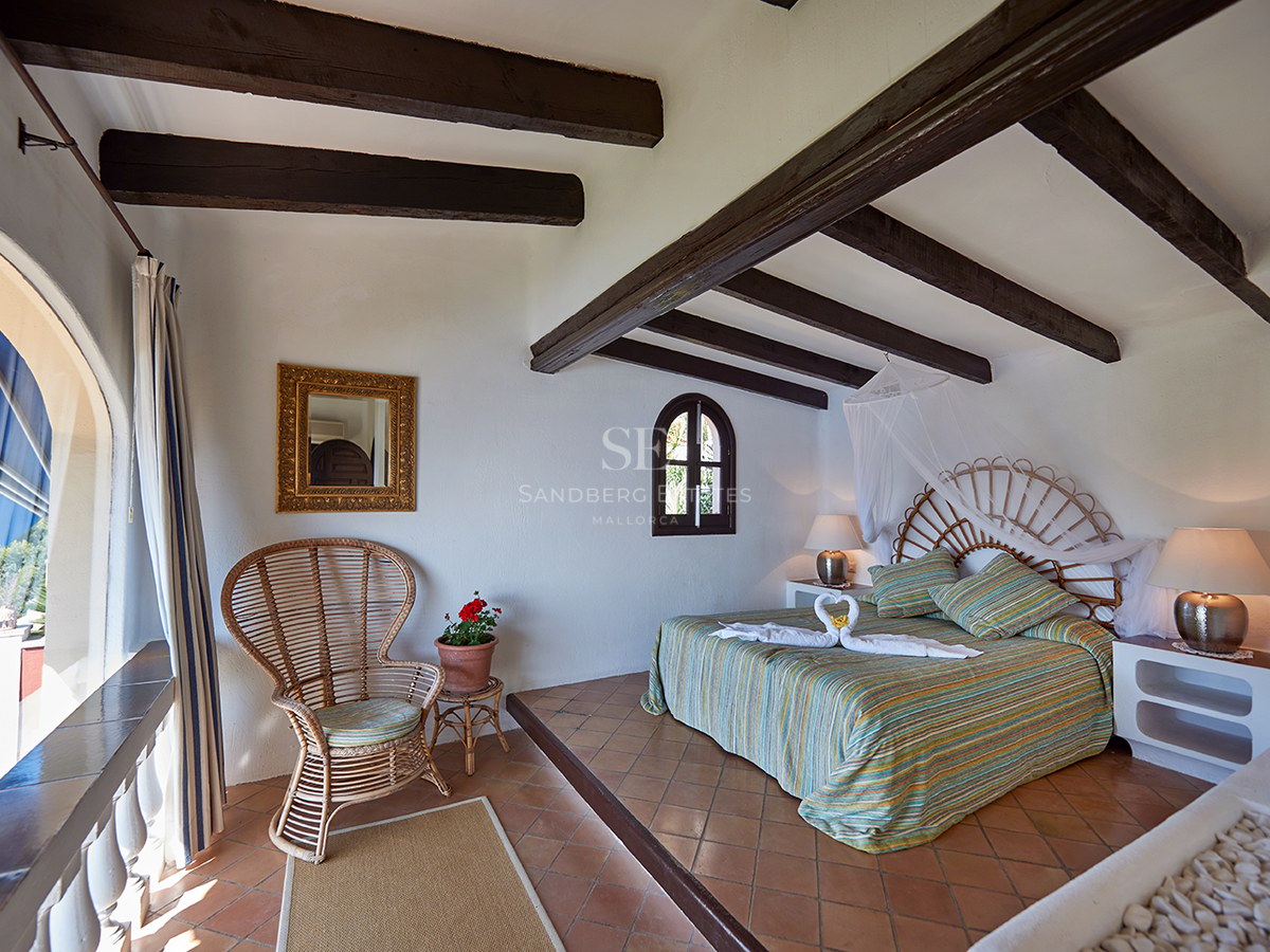 Helles Schlafzimmer mit dunklen Holzbalken, Weidenkopfteil, Terrakottaboden und Rundbogenfenster.