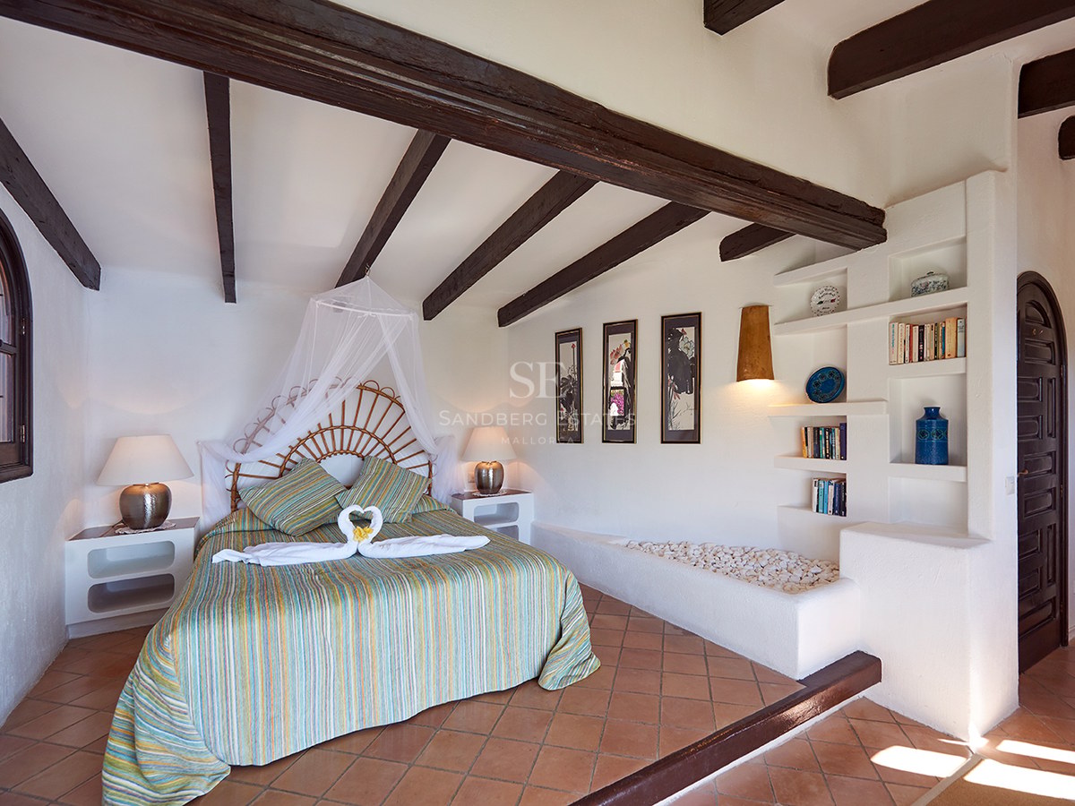 Schlafzimmer mit Korbgeflecht-Bett, dunklen Holzbalken, Terrakottafliesen und weißen Wänden mit eingebauten Regalen.