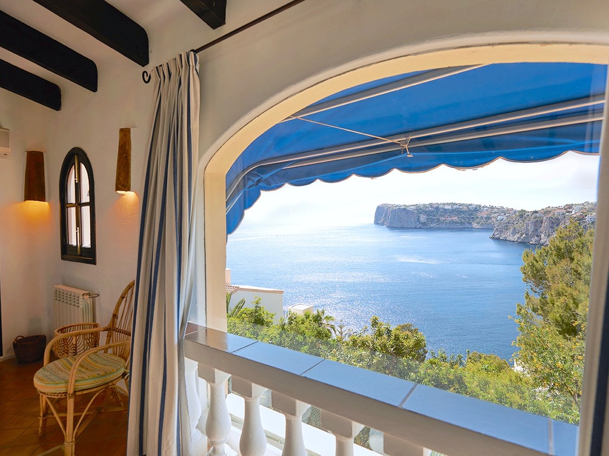 Überdachte Terrasse mit Korbsessel und blauer Markise, mit Blick auf das blaue Meer und Klippen.